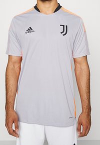 Szary, krótki rękaw, wykonany z lekkiego materiału, z czarnym kołnierzem i logo Adidas. Jasne pomarańczowe akcenty na ramionach. Widoczne logo Juventusu.