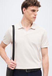 Νεαρός άνδρας που φοράει ανοιχτό μπεζ πόλο πουκάμισο και μπλε παντελόνι, κρατώντας το λουρί ενός μαύρου ώμου τσάντας, κοιτάζει προς τα δεξιά του.