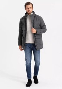 Giubbotto puffer grigio con colletto alto, chiusura a zip e tasche frontali, indossato sopra un maglione chiaro e abbinato a jeans blu e stivali neri.