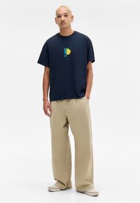 Vyras su trumpais šviesiais plaukais, vilkintis tamsiai mėlyną marškinėlį su mažą spalvingą grafiką, šviesiai rudus plačius kelnes ir šviesiai rudus sportbačius, stovintis prieš baltą fono.