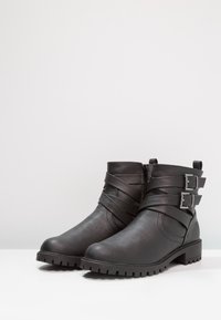Botas negras hasta el tobillo hechas de cuero sintético, con puntera redondeada, detalles de correas horizontales y dos hebillas ajustables en el lateral.