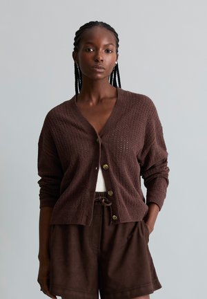 Vrouw met gevlochten haar die een losse bruine gebreide cardigan met knopen draagt over een witte top en bijpassende bruine shorts, staand met één hand in de zak.