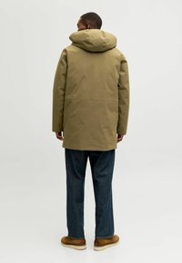 Tamsiai žalios spalvos dideliu gobtuvu atspari vandeniui parka su tiesia kirpimo, šoninėmis kišenėmis. Dėvima ant tamsiai mėlynų džinsų kelnių ir rudų avalynės.