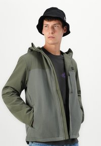 Grøn jakke med hætte, der har lynlås foran, to sidelommer og en glat overflade. Båret over en mørk t-shirt og kombineret med en sort bøttehat.