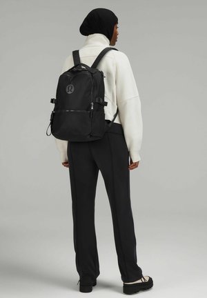 Black backpack with adjustable straps, multiple zippered compartments, and a logo on the front. Worn with a white hoodie and black pants.