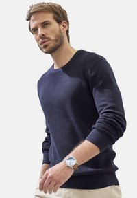 Ein marineblauer gestrickter Pullover mit Rundhalsausschnitt und ribbierten Bündchen. Das Model trägt eine silberne Uhr mit einem hellbraunen Lederarmband. Heller Hintergrund.