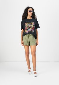 T-shirt graphique noir avec un motif floral multicolore, associé à un short vert olive. Complété par des chaussures blanches et des lunettes de soleil sombres.