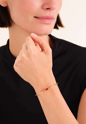 Gouden metalen armband met een dunne, cirkelvormige vorm, voorzien van een accent van een rode edelsteen, gedragen om een pols met een zachte huidtextuur.
