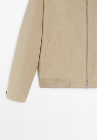 Beżowa długa kurtka z długim rękawem, z pionowym zamkiem z przodu, pionową boczną kieszenią, ściągaczem na dole oraz ozdobnym guzikiem przy mankiecie, na białym tle.