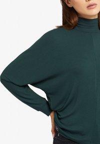 Maglione verde a collo alto con una vestibilità morbida, texture a coste e spacchi laterali, con maniche lunghe e silhouette rilassata.