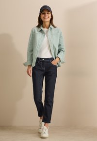 Veste en velours côtelé vert clair sur un t-shirt blanc, associée à un jean en denim foncé et des baskets blanches. Une casquette marine complète l'ensemble.