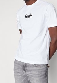 Camiseta blanca de algodón con un logo negro de Calvin Klein en el pecho. Combinada con jeans de mezclilla gris. Diseño simple de cuello redondo.