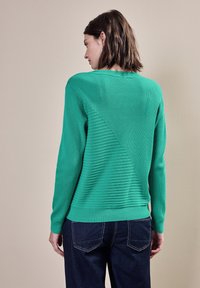 Zelený žebrovaný svetr s diagonálním texturálním detailem na zádech, s dlouhými rukávy a vypasovaným dolním lemem; nošený přes tmavě modré džíny.