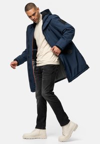 Parka larga azul marino con cremallera frontal, forro de piel y detalles en los hombros. Combinada con un suéter crema, jeans negros y botas blanco roto.