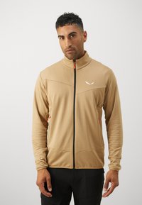 Brun zip-up jakke med teksturert stoff, svart glidelås og oransje glidelåskrok; har et emblem på brystet og sidelommer.