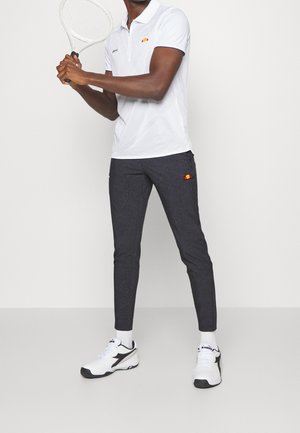 Mężczyzna w białej koszulce polo i ciemnych spodniach sportowych trzymający rakietę tenisową, ubrany w białe buty sportowe, stoi na jednolitym tle.