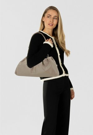 Mujer con un cárdigan negro con ribete blanco, pantalones negros, llevando un gran bolso de hombro beige con detalles fruncidos.
