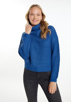 Pulover albastru cu guler înalt, tricotat, cu un model texturat, cu o croială supradimensionată și manșete cu nervuri. Purtați cu blugi închiși la culoare pe un fundal deschis.
