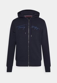 Marineblauwe zip-up hoodie met zijzakken, capuchon met trekkoord en de handtekening "Tommy Hilfiger" in blauwe script letters over de borst.