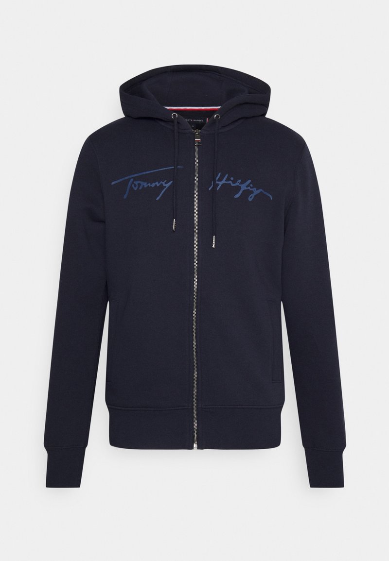 Marineblauwe zip-up hoodie met zijzakken, capuchon met trekkoord en de handtekening "Tommy Hilfiger" in blauwe script letters over de borst.