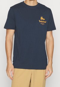 Námořnická modrá bavlněná tričko s logem Timberland a grafikou boty v žluté barvě na hrudi. Krátké rukávy a design s kulatým výstřihem.