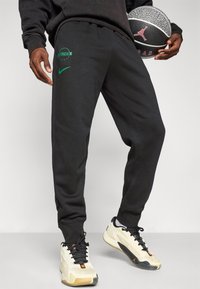 Osoba oblečená v čiernych Nike teplákových nohaviciach Celtics a béžových teniskách, ktorá drží čierno-biely basketbal Jordan.