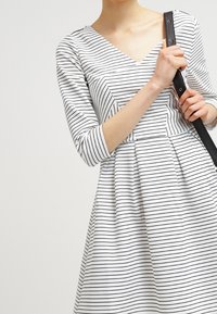 Kobieta w białej sukience z cienkimi czarnymi poziomymi paskami i rękawami sięgającymi do trzeciej części przedramienia, trzymająca czarny pasek torebki na ramię.