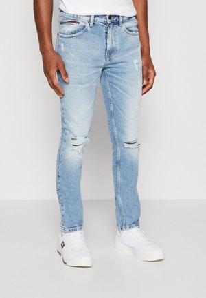 Mężczyzna w jasnoniebieskich podartych dżinsach, białych trampkach i białej koszulce, stojący na jednolitym białym tle.