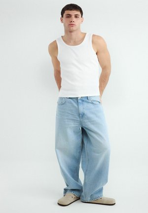 Mladý muž stojí s rukami za chrbtom, nosí biele tielko, široké svetlomodré džínsy a béžové slip-on topánky na jednofarebnom pozadí.