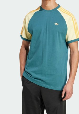 Mężczyzna w turkusowo-kremowej koszulce Adidas z żółtymi paskami na rękawach i czarnych spodniach, stojący z lewą ręką widoczną.