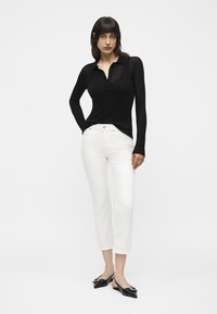 Woman standing with hands in pockets wearing a black long-sleeve top, white cropped pants, and black pointed-toe flats.
