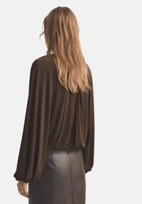 Blusa marrón de manga larga con espalda drapeada, hecha de tela suave. Combinada con una falda de cuero marrón oscuro, con puños elásticos.