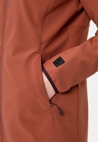 Hand in pocket of rust-colored jacket with stitched seam details, visible zipper pull, and black fabric label on sleeve.