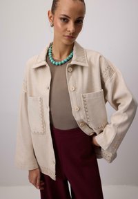 Beżowa kurtka z mieszanki wełny, zdobiona misterną haftowaną dekoracją, o luźnym kroju, z dwoma przednimi kieszeniami i beżowymi guzikami. Noszona z brązową bluzką i bordowymi spodniami.