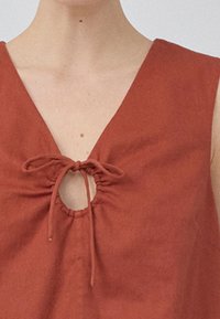 Haut sans manches de couleur rouille avec un col en V et un cordon central, créant une ouverture en forme de clé. Tissu doux et texturé.