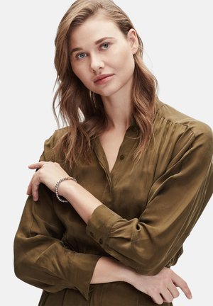 Olijfgroene shirt met knoopmanchetten; model met een zilveren kralenarmband om de pols. Zachte, gestructureerde stof met een subtiele glans.