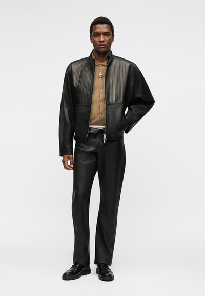 Άνδρας με μαύρο δερμάτινο μπουφάν με κατακόρυφη ραφή, μαύρο δερμάτινο παντελόνι, καφέ πουκάμισο με κουμπιά, μαύρα παπούτσια και περιδέραιο με μενταγιόν.