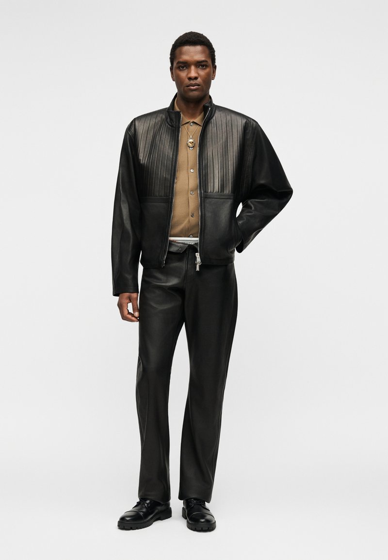 Homme portant une veste en cuir noire avec des coutures verticales, un pantalon en cuir noir, une chemise marron boutonnée, des chaussures noires et un collier pendentif.