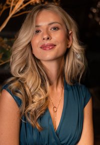 Cheveux blonds coiffés en douces vagues, portant une robe bleu sarcelle. Un délicat collier en or avec un pendentif circulaire repose sur sa clavicule.