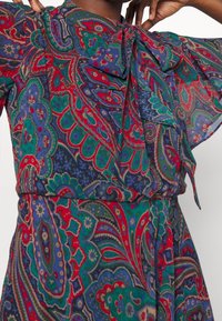 Färgglad klänning med paisleymönster, försedd med knytkrage, korta utsvängda ärmar och midjebälte. Materialen verkar lätta och luftiga.