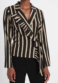 Blazer portefeuille rayé noir et beige avec bordure à volants, encolure en V et détail à nouer, confectionné dans un tissu lisse avec une coupe ajustée.