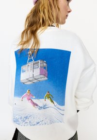 White sweatshirt featuring a colorful graphic of skiers against a blue sky, with a cable car above labeled "APRES SEASON."