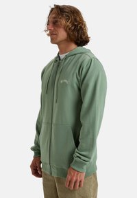 Hoodie zippé vert avec cordons de serrage, doté d'une poche kangourou et d'un logo brodé. Fabriqué en tissu doux et texturé avec une coupe ajustée.