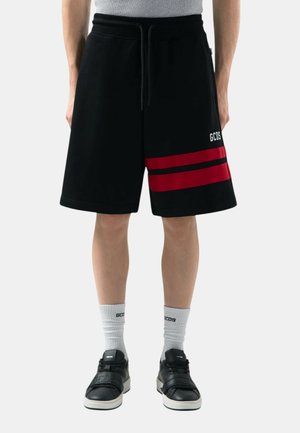 Sorte shorts med røde og sorte vandrette striber på det ene ben, båret med hvide GCDS-strømper og sorte sneakers på en person, der står.