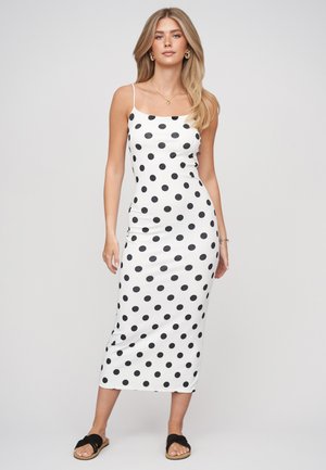 Donna che indossa un vestito midi bianco con pois neri, spalline sottili e sandali neri a ciabatta, in piedi davanti a uno sfondo neutro.
