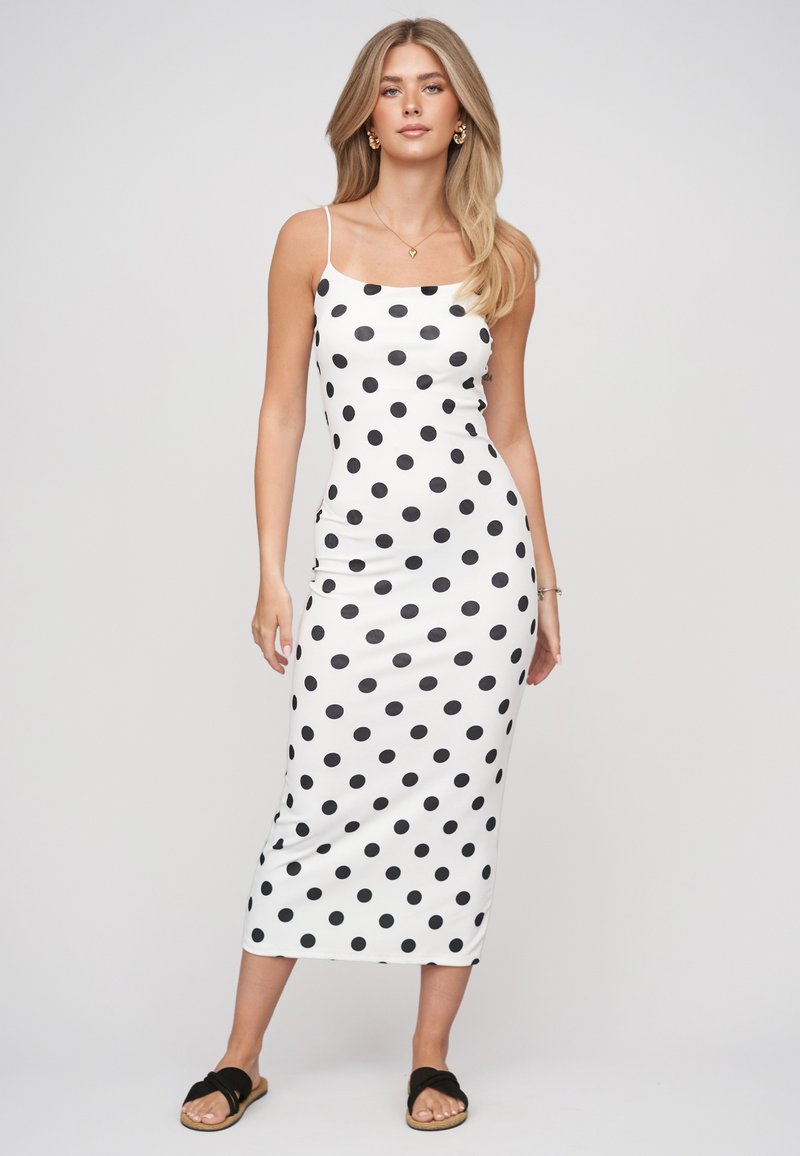 Femme portant une robe midi blanche à pois noirs, fines bretelles et sandales slides noires, debout devant un fond neutre.