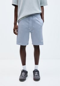Shorts en sweat bleu clair avec taille élastique, poches latérales et détail de logo. Associés à un t-shirt gris ample et des baskets noires.