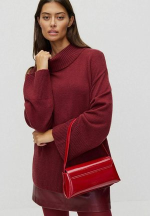 Pull bourgogne avec un col haut à côtes, associé à un sac à main rouge brillant. Le pull a des manches surdimensionnées. Design épuré et moderne.