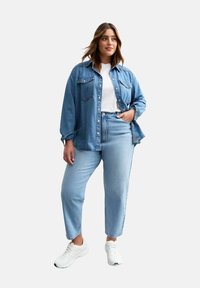 Camisa de denim en azul claro, con dos bolsillos en el pecho y botones, sobre una camiseta blanca. Jeans rectos en azul claro y zapatillas blancas completan el look.