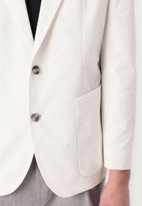 White textured blazer with two black round buttons, a chest pocket, and notched lapels. Fabric appears lightweight and smooth.